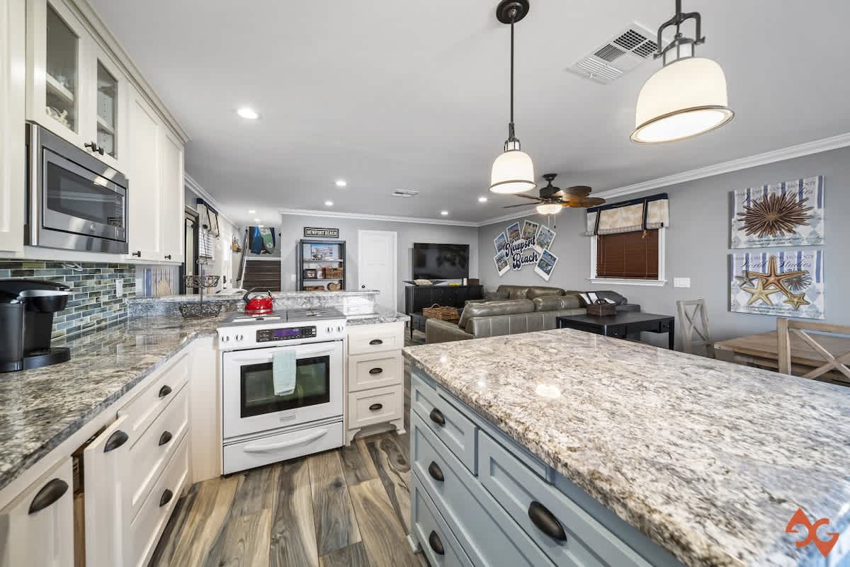 Modern kitchen with light granite countertops, white shaker cabinets, a white electric range, and a blue-grey island, opening into a living area with a grey sofa and wall-mounted TV.