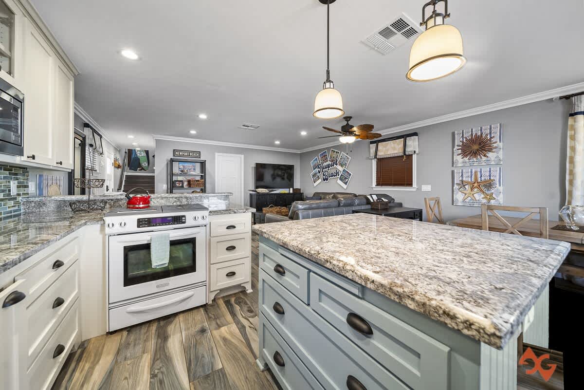 Contemporary kitchen with a light blue island and granite countertops, overlooking an open living area with wood-look flooring and a ceiling fan.