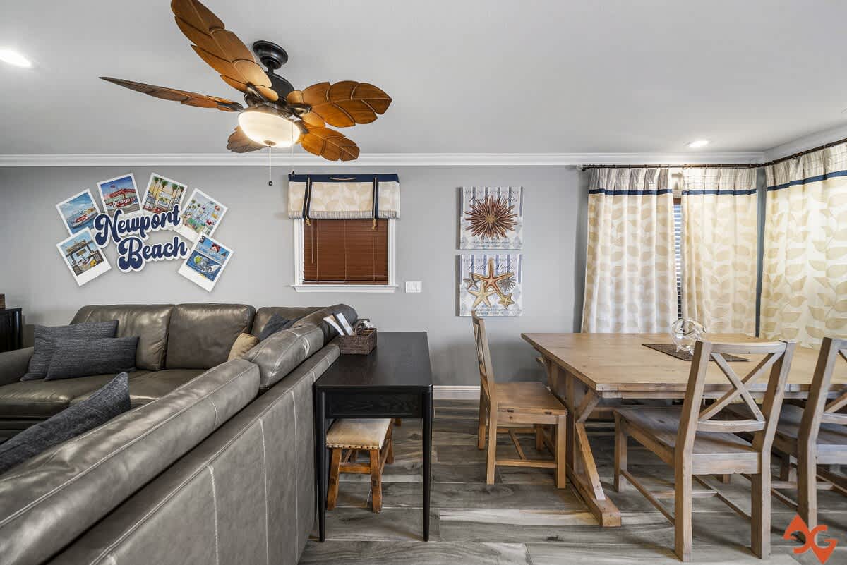 Bright dining area with a rustic wooden table and matching chairs, situated near a window with patterned curtains and a ceiling fan in the adjacent living space.