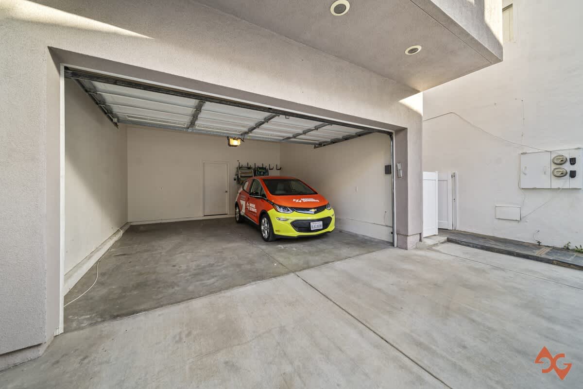A spacious, well-lit garage with an open door reveals a bright orange and yellow car parked inside, featuring a clean concrete floor and white walls.