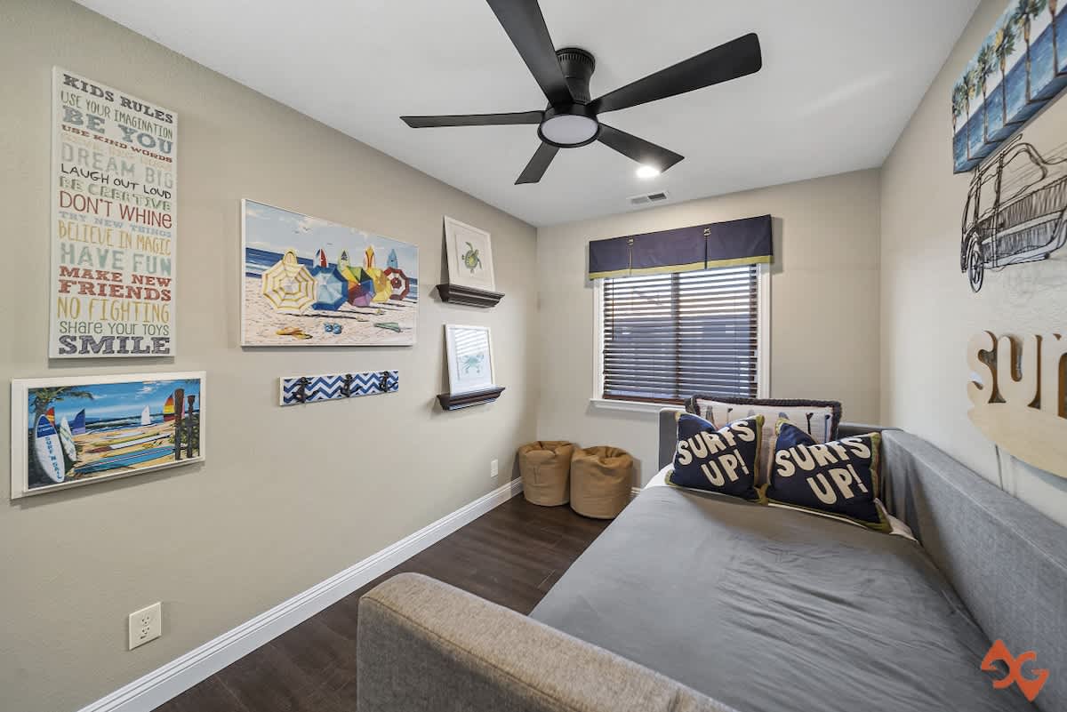 Bright kids' bedroom with a gray daybed, dark wood floors, and a window with blinds, adorned with colorful beach-themed artwork and 'Kids Rules' wall decor.