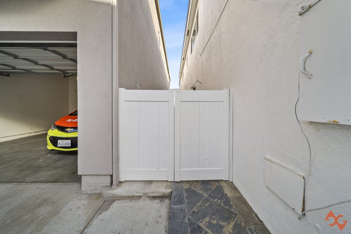 A narrow concrete and stone-tiled walkway between two buildings features a white privacy gate, with a partial view of a bright car parked inside an open garage on the left.