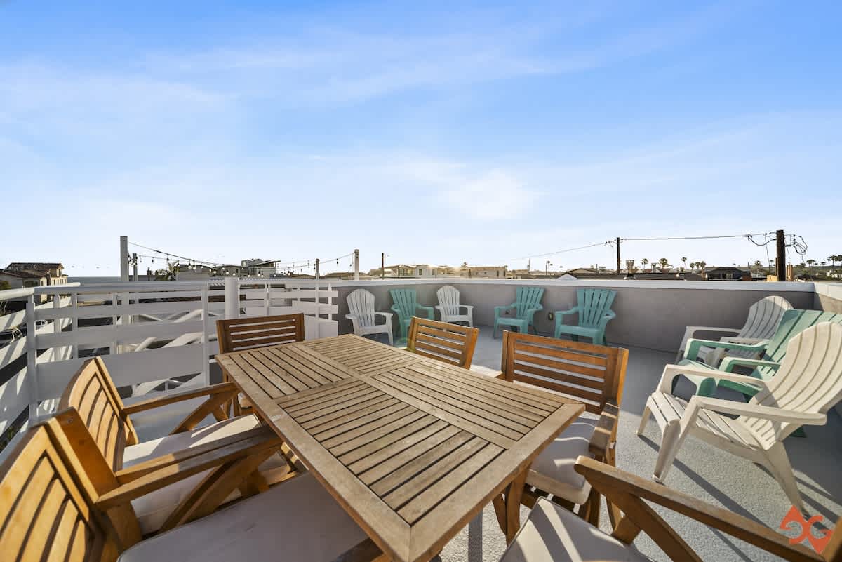 An expansive rooftop deck features a wooden outdoor dining table with chairs on one side and a collection of light green and white Adirondack chairs on the other, all enclosed by a white railing with panoramic views.