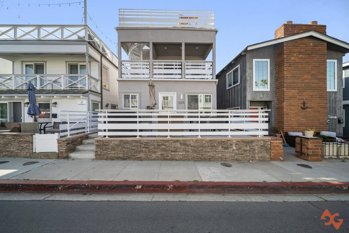 A wider view of the three-story house's front exterior, showcasing its light gray stucco facade, white horizontal slatted fence on a stone base, and the neighboring properties on either side.