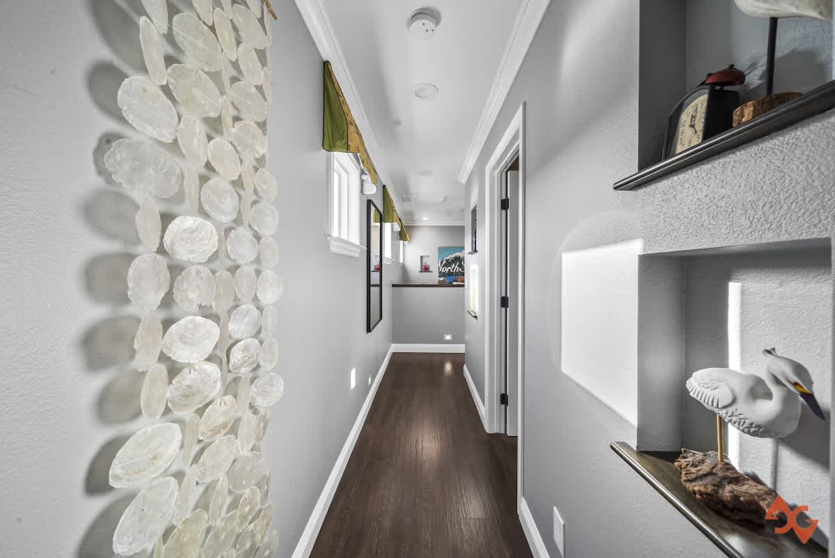 Narrow hallway with light grey walls, dark wood flooring, and decorative wall art made of shell-like discs, leading towards a mirror and another room.
