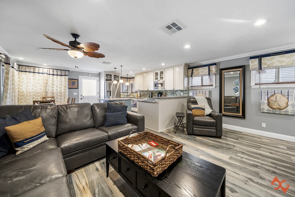 Inviting living room with a dark gray leather sectional sofa, a black coffee table, and wood-look flooring, opening to a modern kitchen with white cabinetry and a breakfast bar.