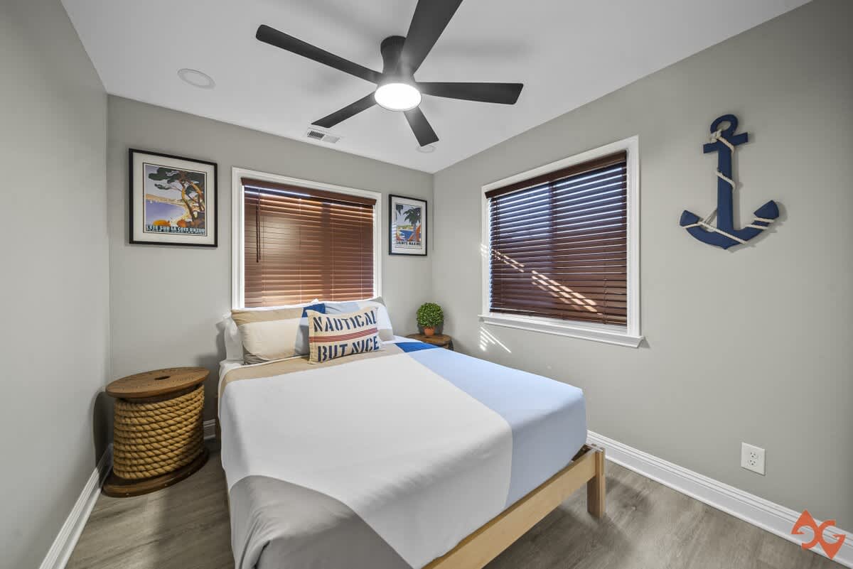 A cozy bedroom with light gray walls, a queen-sized bed with blue and white bedding, and two windows with dark wood blinds. A black ceiling fan with a light fixture hangs overhead, and a large blue anchor decorates the wall.