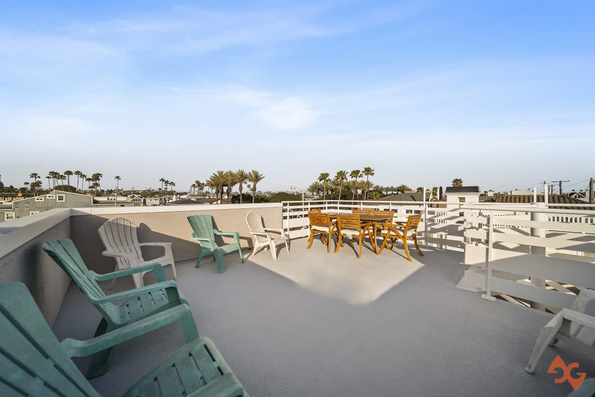 An expansive rooftop deck with a mix of light green and white Adirondack chairs and a wooden dining table with chairs, offering panoramic views of the surrounding neighborhood and distant palm trees under a clear blue sky.
