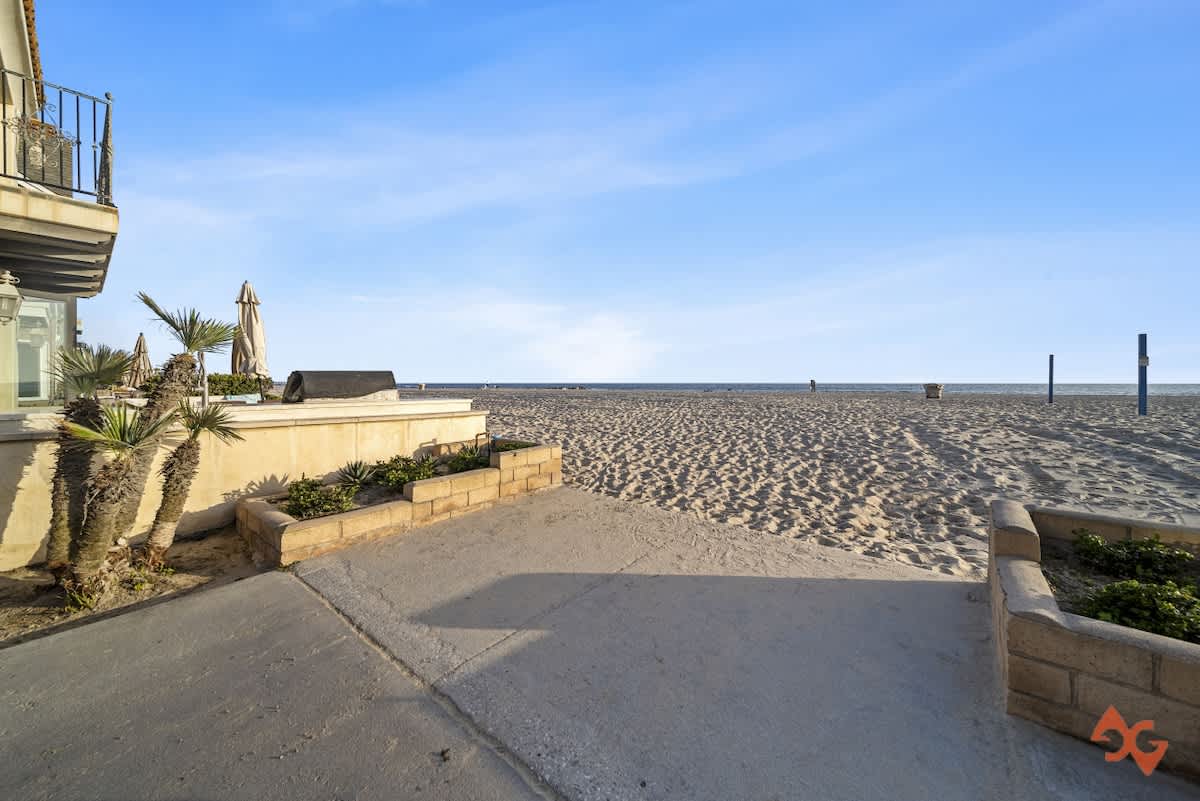 A concrete pathway leads directly to a wide, sandy beach under a clear blue sky, with the ocean visible in the distance and a building with a small balcony and palm trees to the left.