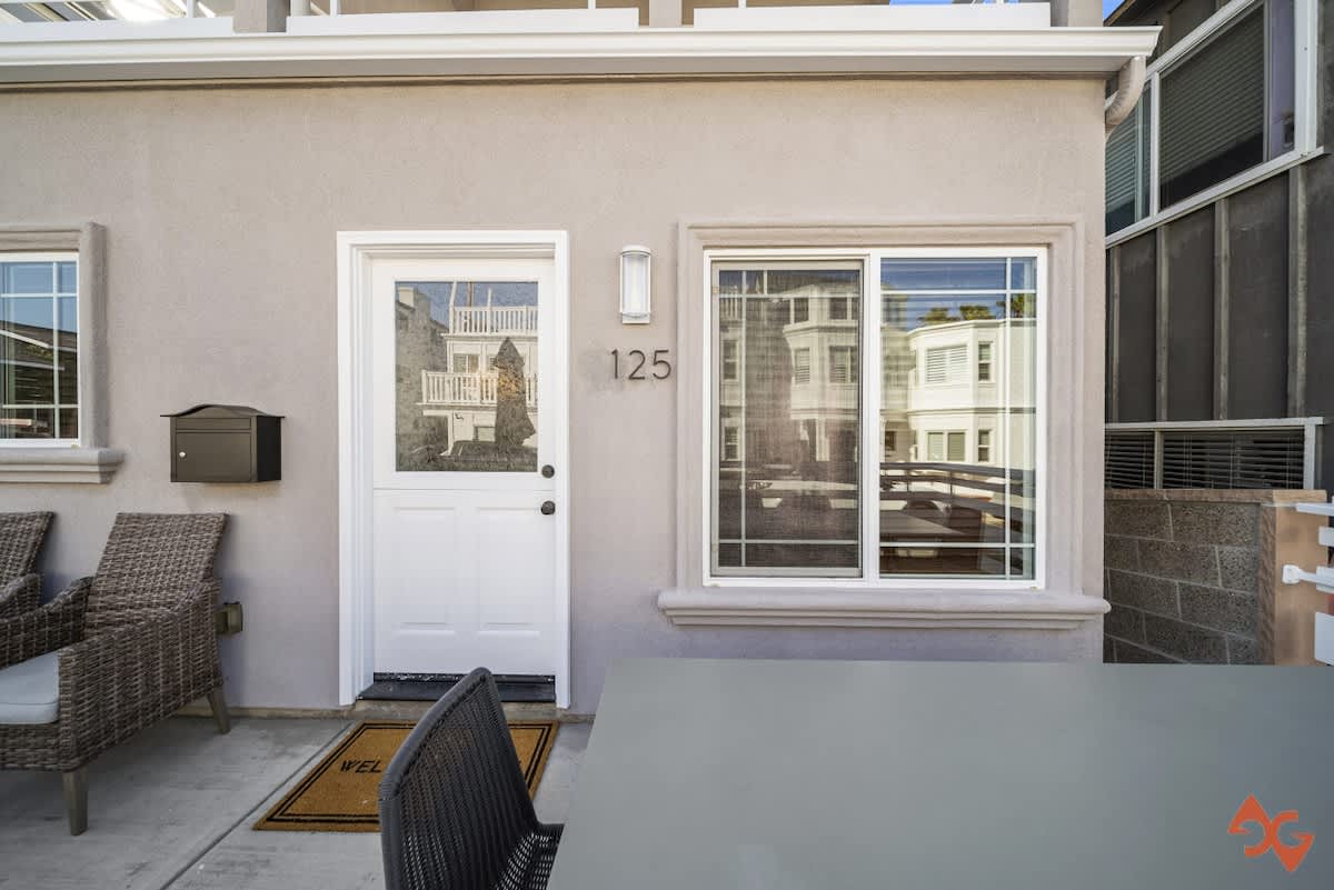 A close-up of the house's front entrance features a white door with a window, a black mailbox, and the house number "125" mounted on the light gray stucco wall, with an outdoor table visible in the foreground.