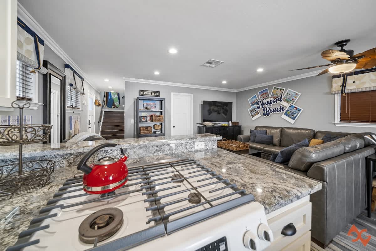 Kitchen counter with a white gas stove and red kettle, offering a view into the open living room with a large gray sectional sofa and wall decor.