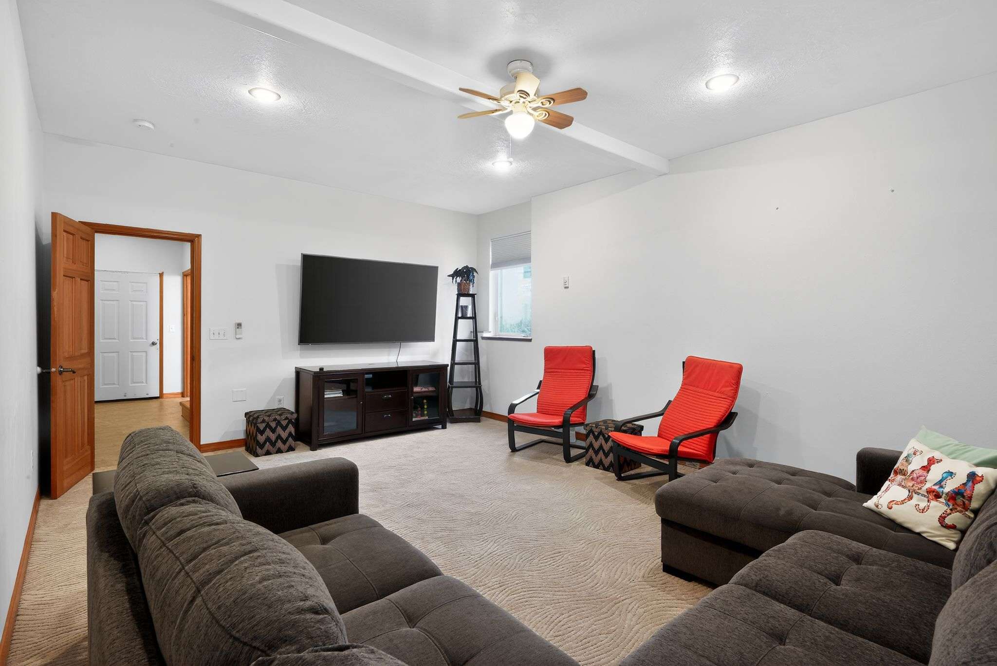 A bright living room with light patterned carpet, white walls, a large dark sectional sofa, and a big screen TV on a dark media console, complemented by two modern red chairs and a ceiling fan.
