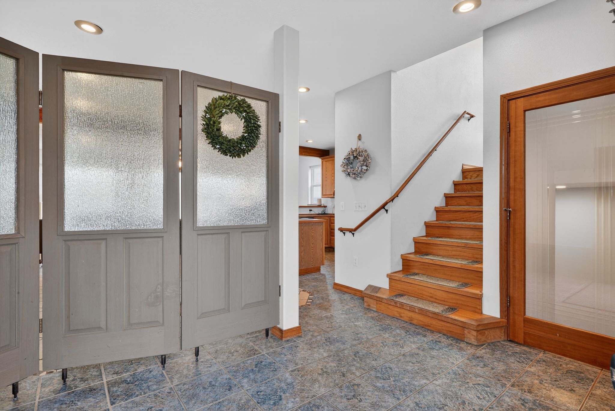 A welcoming foyer with blue patterned tile flooring, a wooden staircase with a railing, and a set of frosted glass saloon-style doors adorned with a wreath, leading to other areas of the house.