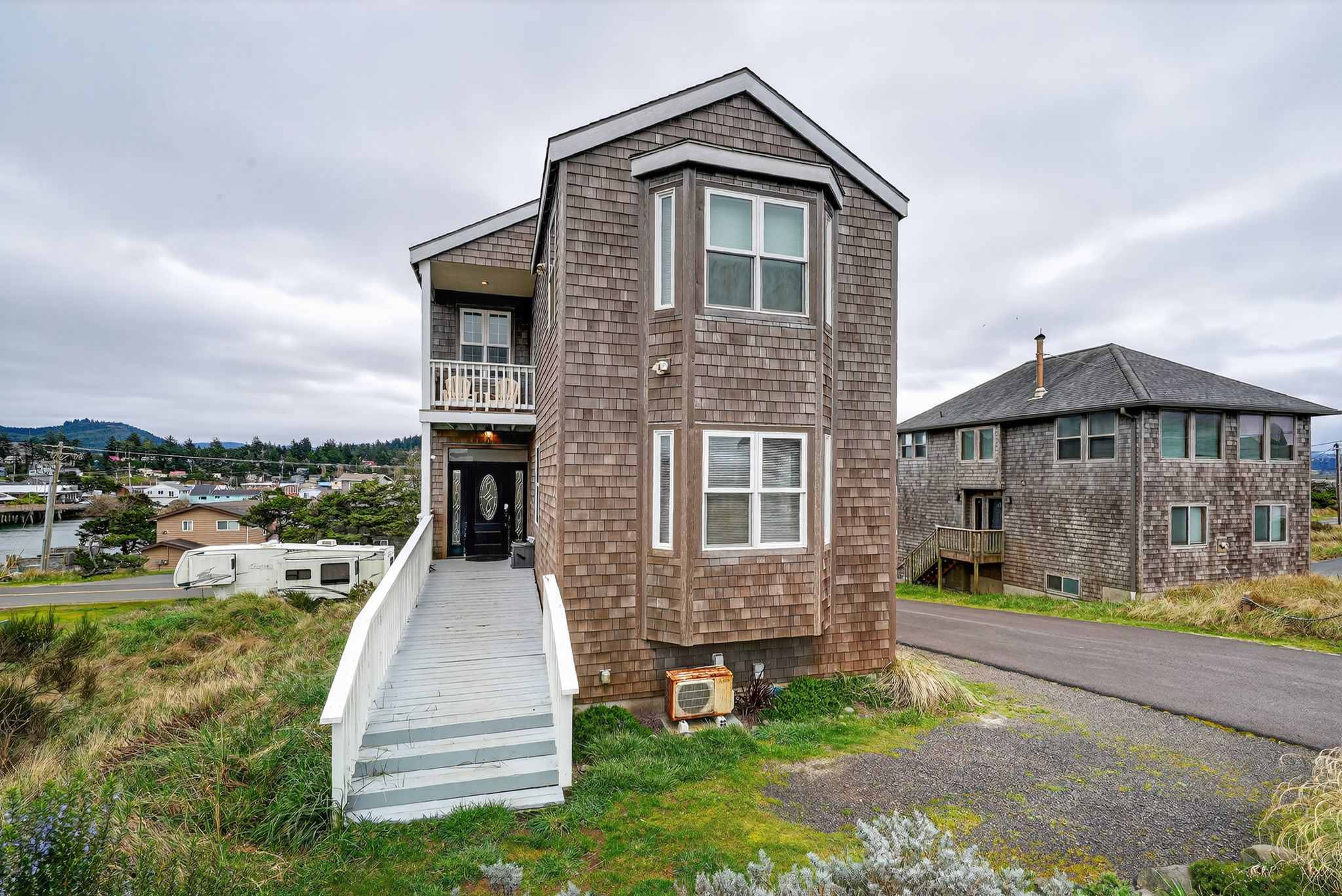 The front and side of the tall, narrow cedar shake house are visible, featuring multiple windows and a prominent wooden ramp leading to the entrance. A small gravel driveway and grassy areas surround the property, with a town in the background.
