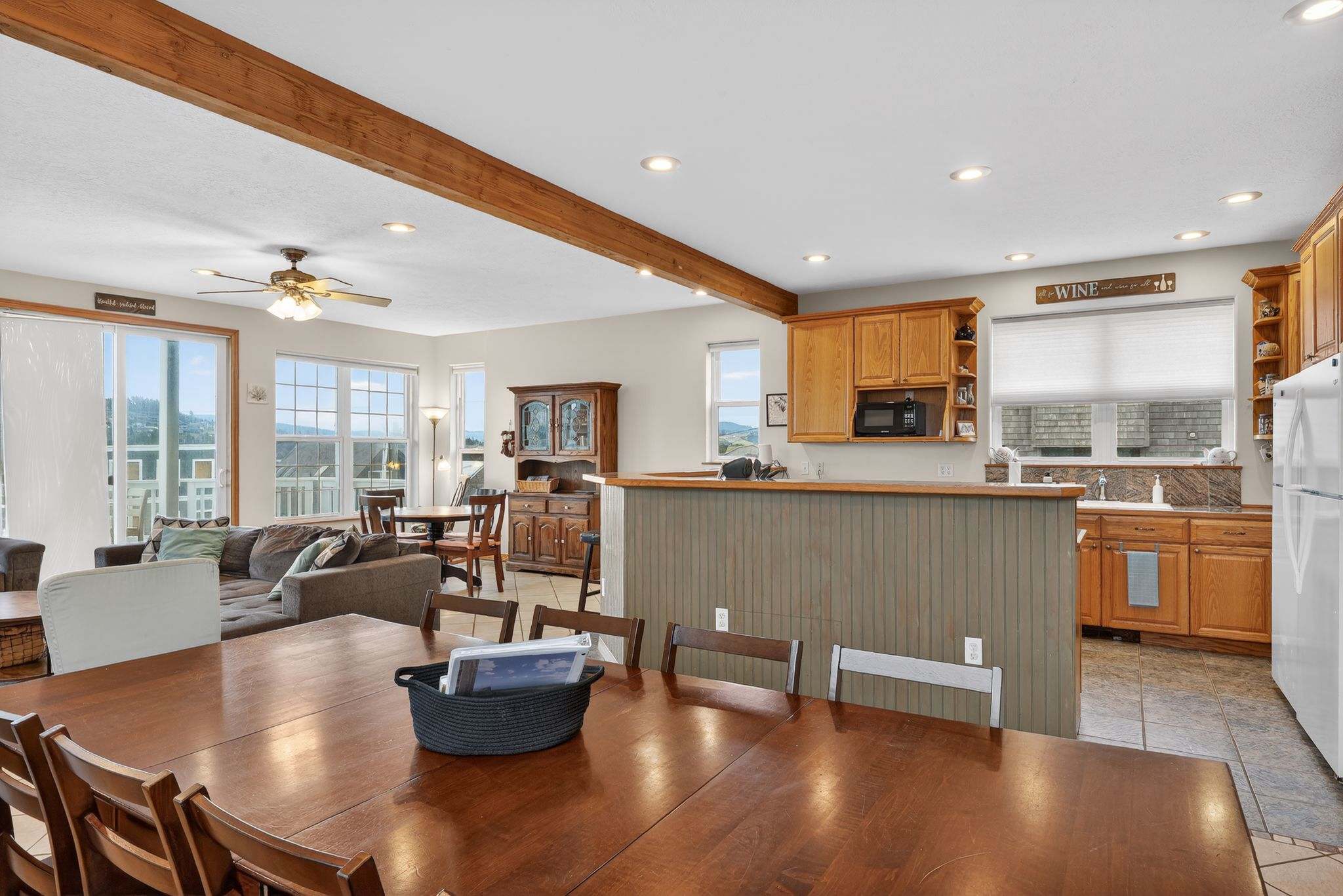 This expansive open-concept area features a large wooden dining table with chairs, adjacent to a kitchen bar with beadboard paneling. The space seamlessly connects to the living room, all under recessed lighting and a prominent wooden beam.