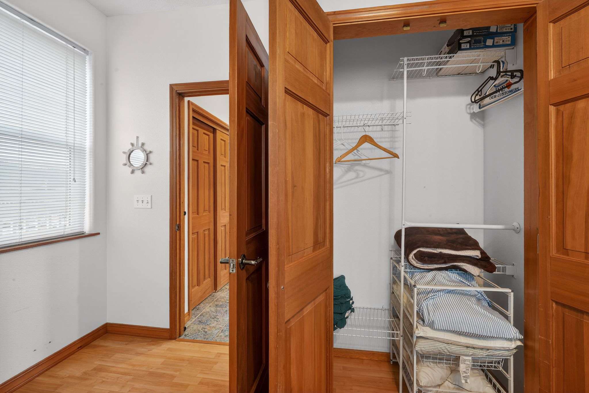 A hallway with light wood flooring and white walls leads to an open wooden closet door, revealing wire shelving stocked with linens and hangers. Another wooden door is visible further down the hallway, suggesting additional rooms.