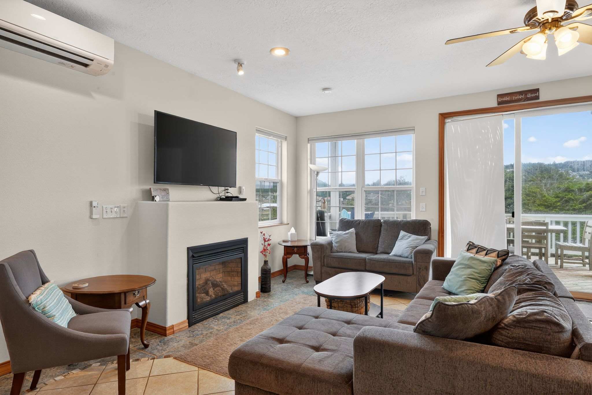 The inviting living room showcases a large brown sectional sofa and a smaller gray sofa, arranged around a modern fireplace with a wall-mounted TV. Expansive windows provide abundant natural light and scenic views, complementing the tile flooring and area rug.