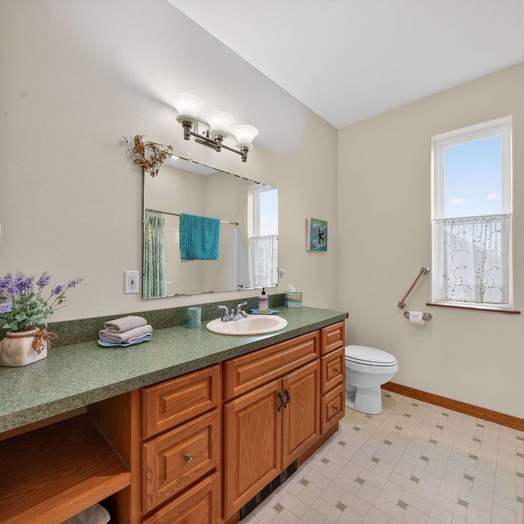 This light-colored bathroom showcases a wooden vanity with a green speckled countertop and a single sink, beneath a mirror. A toilet is positioned next to a window adorned with sheer curtains, and the floor features light-colored patterned tiles.