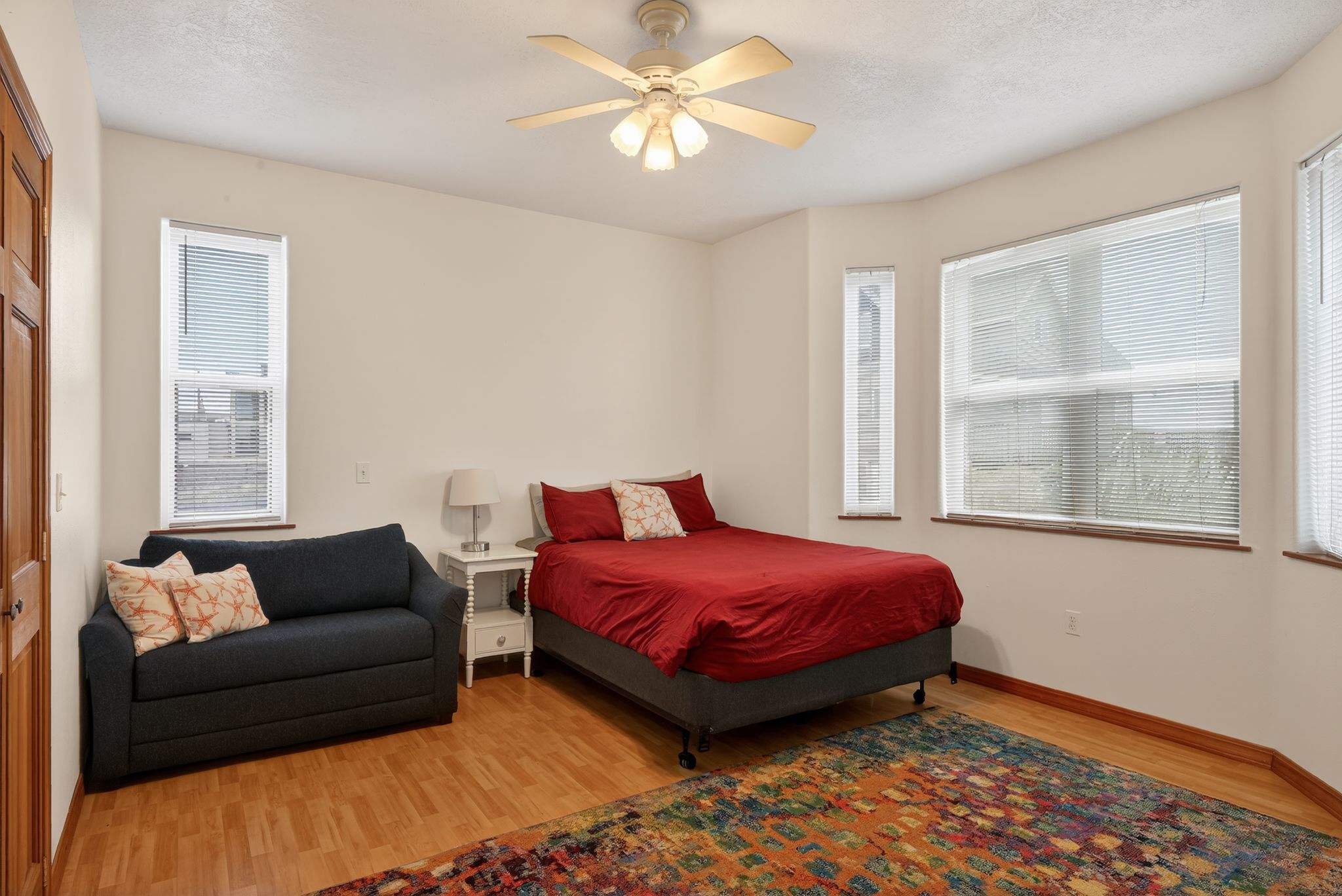 A bright bedroom with light walls and hardwood floors features a bed with red bedding and a dark gray sofa. A white nightstand with a lamp is between the bed and sofa, and a colorful rug adds warmth.