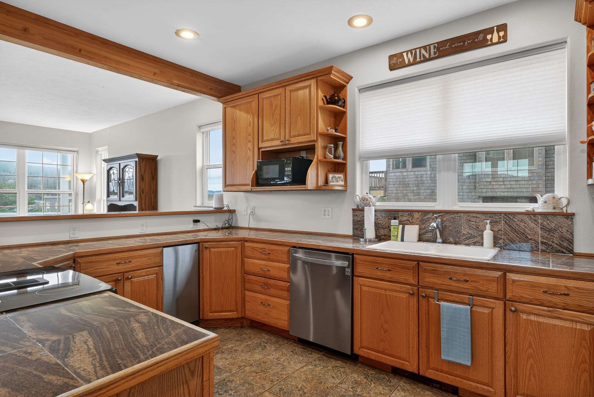 A bright kitchen area highlights wooden cabinetry, dark granite-look countertops, and a white sink with a window above it. Stainless steel appliances, including a dishwasher, are integrated into the design, with tile flooring throughout.