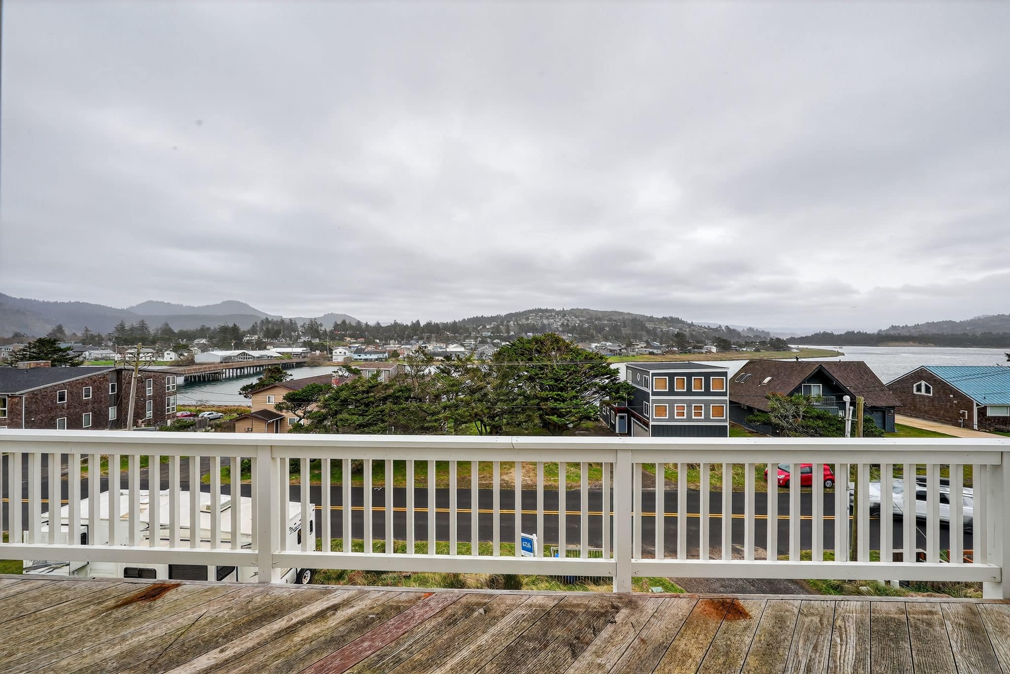 A wide shot from a wooden deck with white railings, offering a panoramic view of a coastal town, a bridge, and a body of water under an overcast sky.