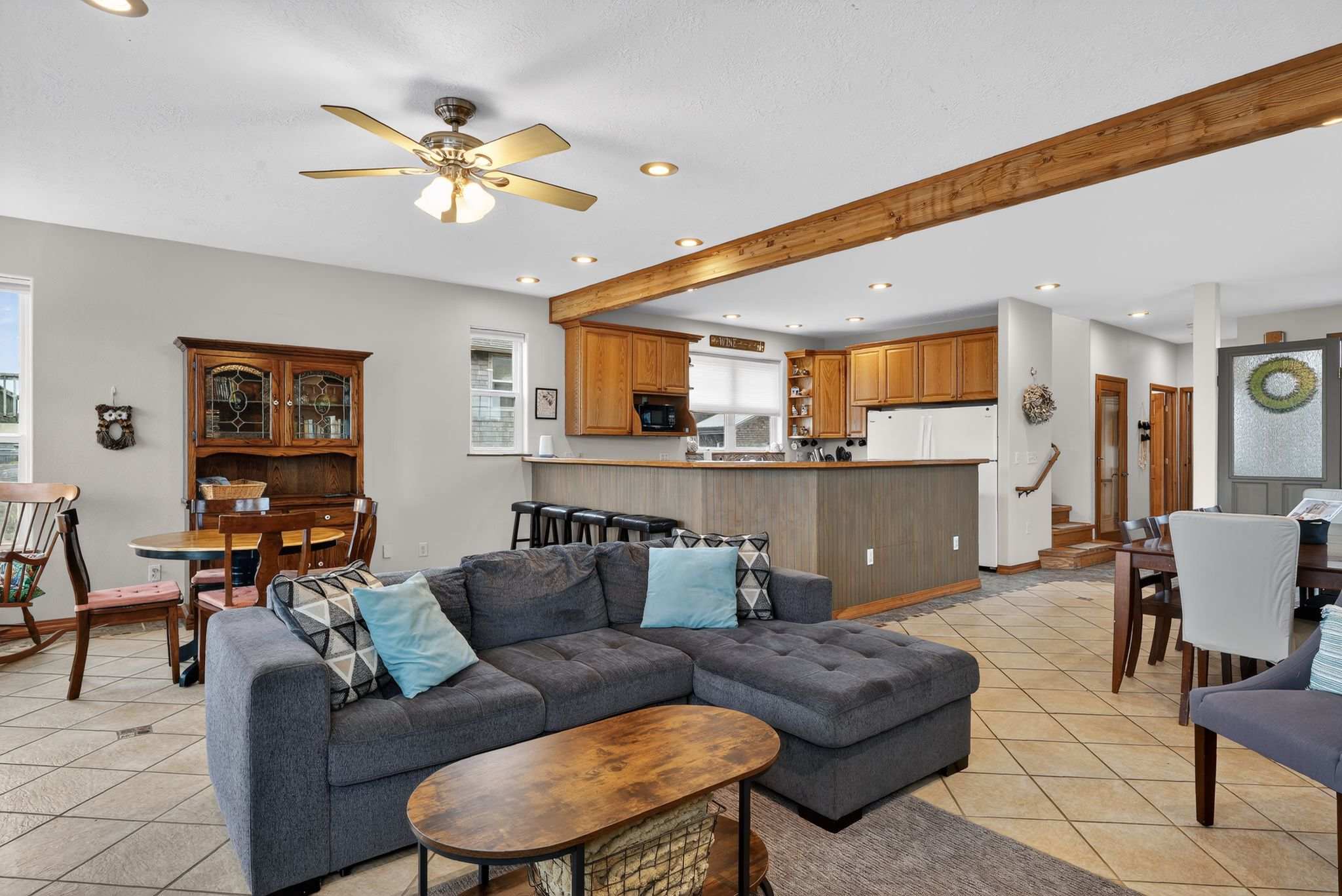 An open-concept living area with a large gray sectional sofa and a wooden coffee table on a rug, flowing into a kitchen with wooden cabinets and a breakfast bar, and a dining area, all featuring tile flooring and a ceiling fan.