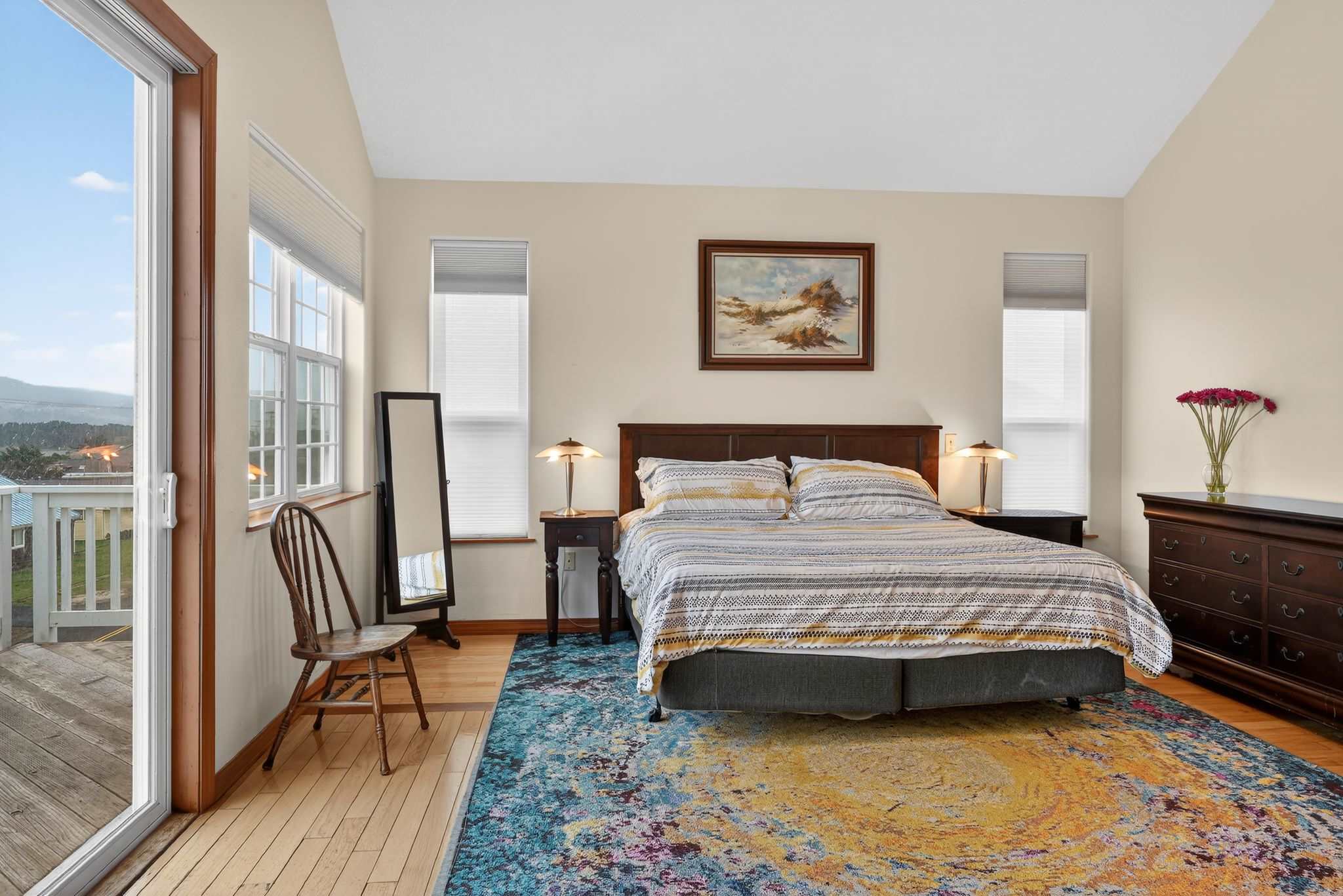 Bedroom with hardwood floors and a colorful abstract rug, featuring a large bed with striped bedding, two nightstands with lamps, a full-length mirror, and a sliding glass door opening to a balcony with ocean views.