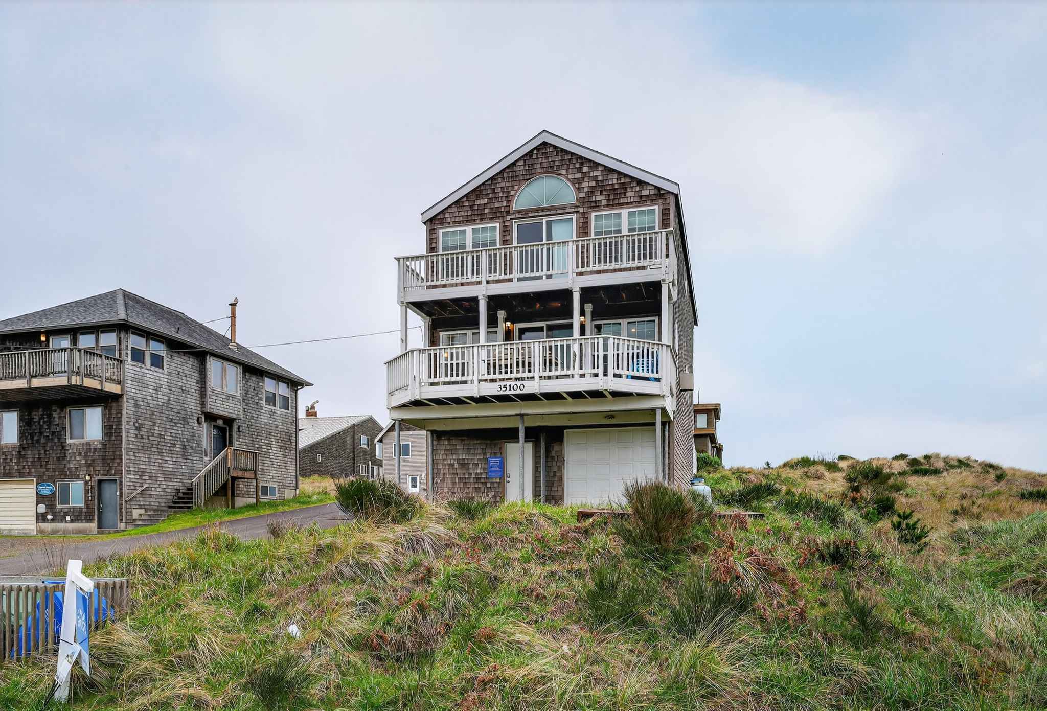 The three-story cedar shake house stands tall on a grassy dune, featuring two balconies with white railings and a ground-level garage. The address "35100" is clearly visible on the lower balcony.