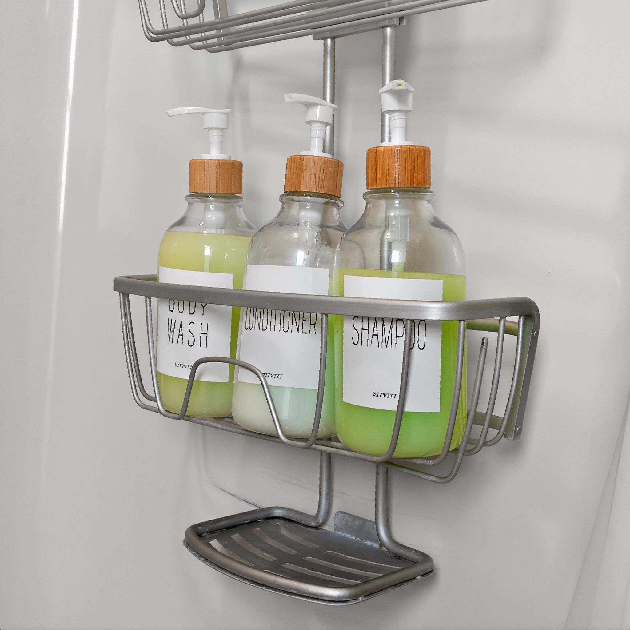 A close-up view captures a metal shower caddy holding three clear bottles labeled 'BODY WASH', 'CONDITIONER', and 'SHAMPOO', each featuring a pump dispenser and a bamboo-style top.