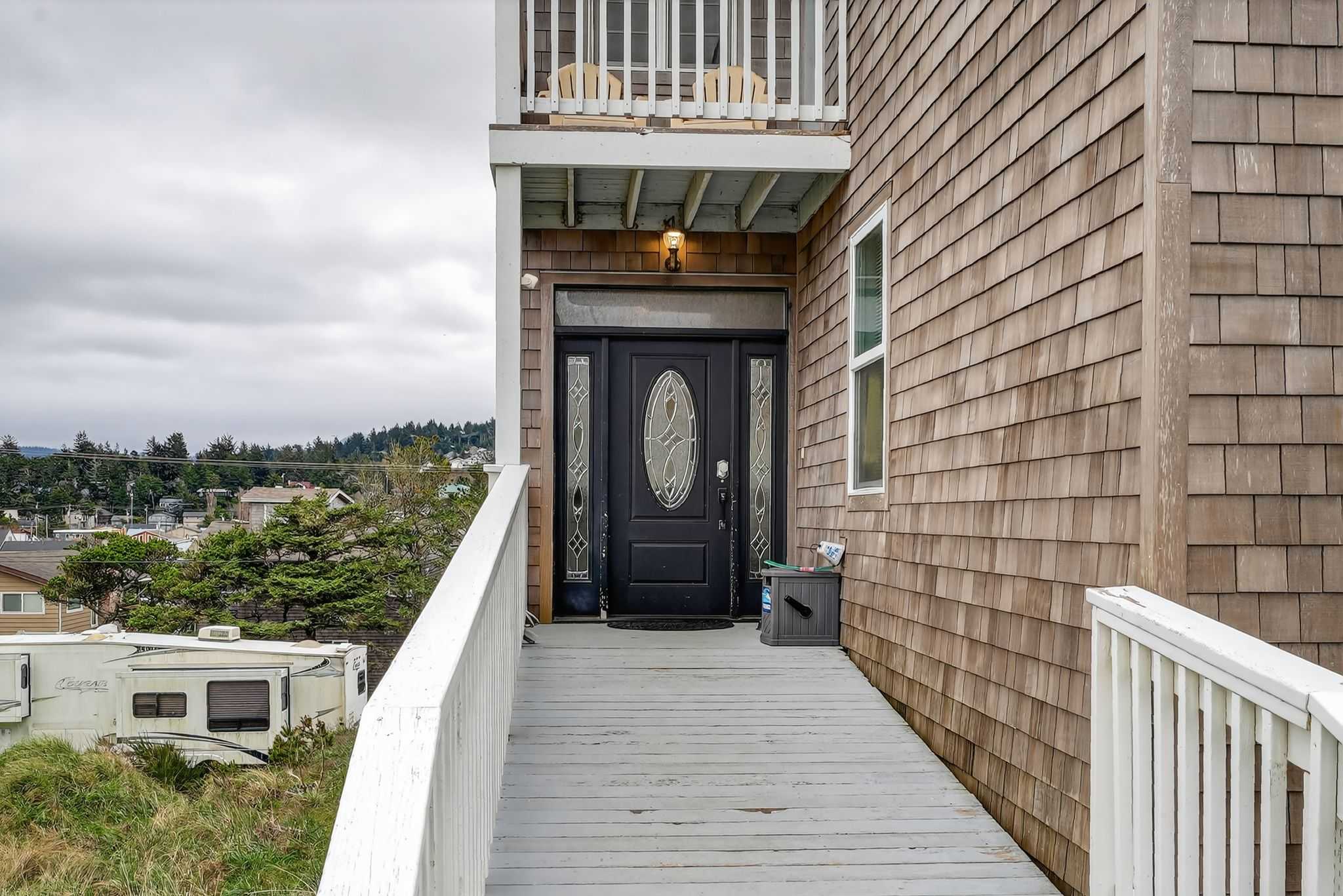 A light grey wooden ramp with white railings leads up to the dark front door of the cedar shake house. A small RV is parked nearby, and distant houses are visible under a cloudy sky.
