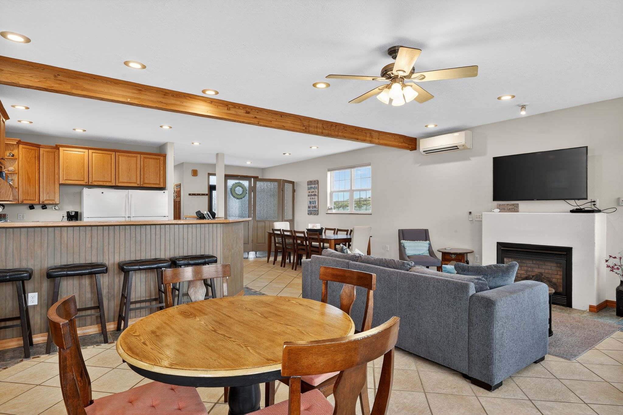 The kitchen features a breakfast bar with dark stools and a small round wooden dining table with chairs in the foreground. Beyond, the open-concept space reveals the living room with a sofa and fireplace, all on tile flooring.