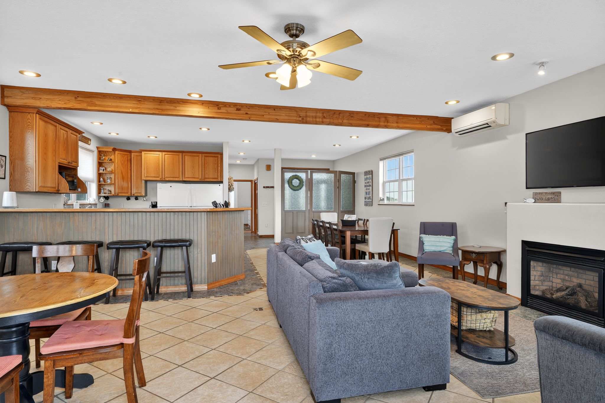 An open-concept living area features a gray sofa facing a fireplace with a wall-mounted TV above it, separated from the kitchen and dining by a prominent wooden beam. Tile flooring extends throughout the space, with large windows providing natural light.