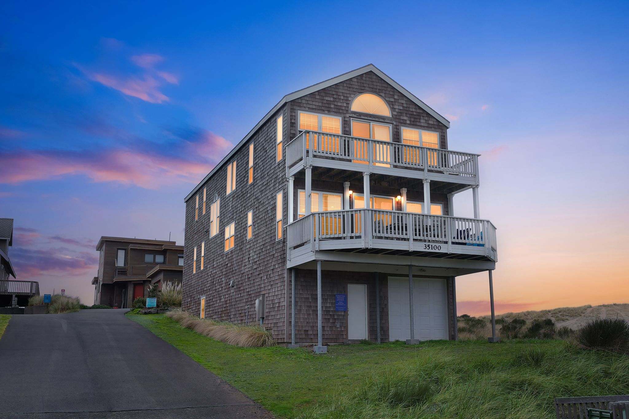 A multi-story shingle-sided house with multiple balconies and white railings, situated on a grassy dune with a paved driveway, under a dramatic sunset sky.
