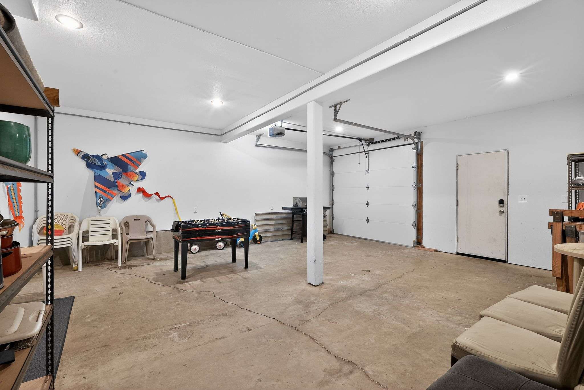 A well-lit garage with a concrete floor and white walls, showcasing a foosball table, various storage items, and a large garage door, with additional seating visible on the right.