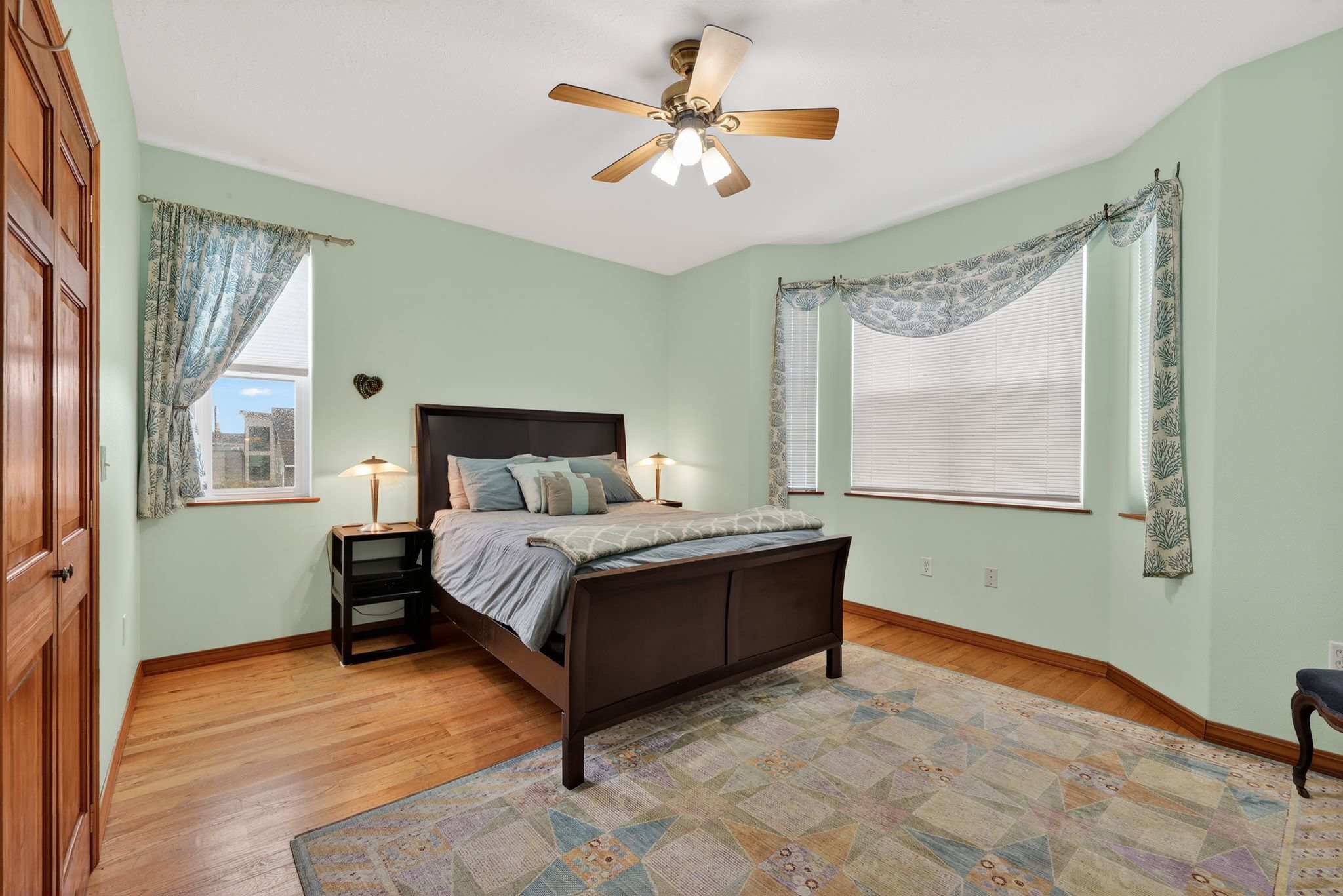 A bedroom with light mint green walls and hardwood floors features a dark wooden bed with blue bedding, flanked by nightstands. Bay windows with blinds and patterned valances offer natural light, and a ceiling fan is overhead.
