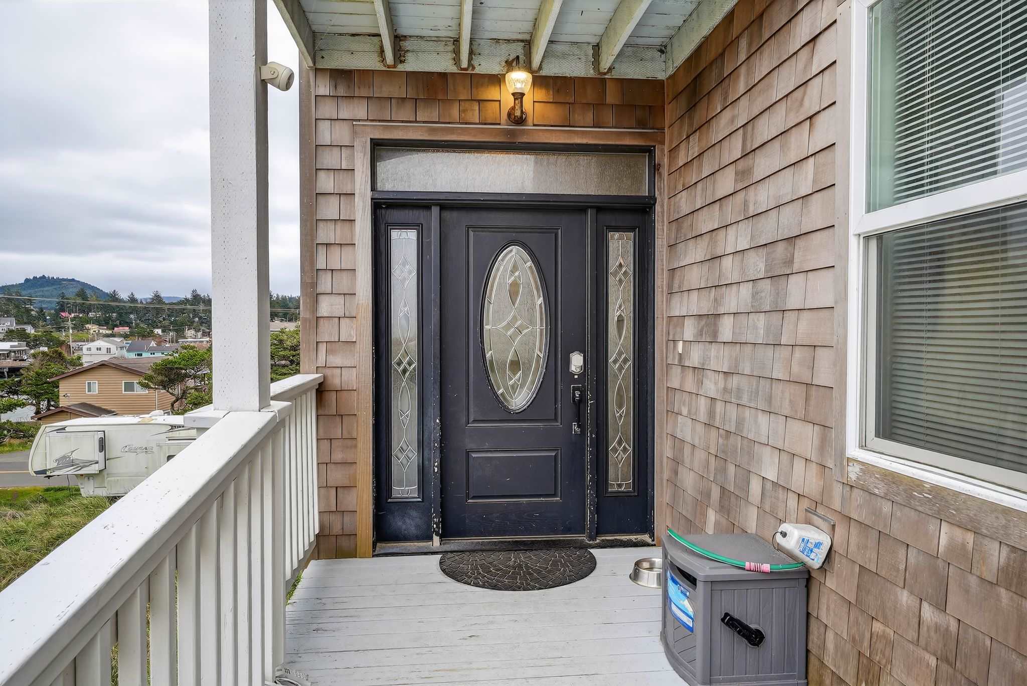 The main entrance features a dark door with an oval glass insert and decorative side panels, set into a cedar shake wall. A black doormat, a pet bowl, and a storage box are visible on the light grey wooden porch floor.