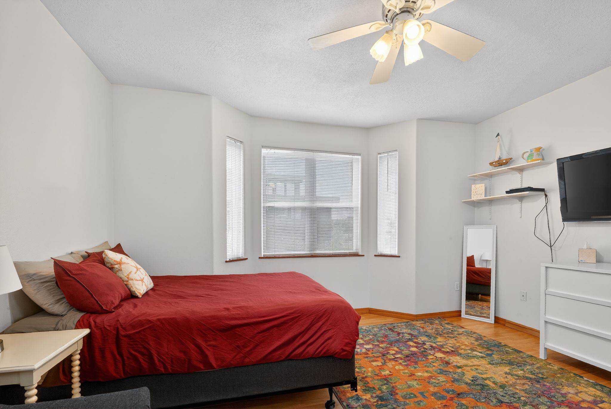 A bright bedroom with light walls and hardwood floors features a bed with red bedding and a bay window with blinds. A white dresser with a TV sits opposite the bed, and a colorful rug covers the floor.