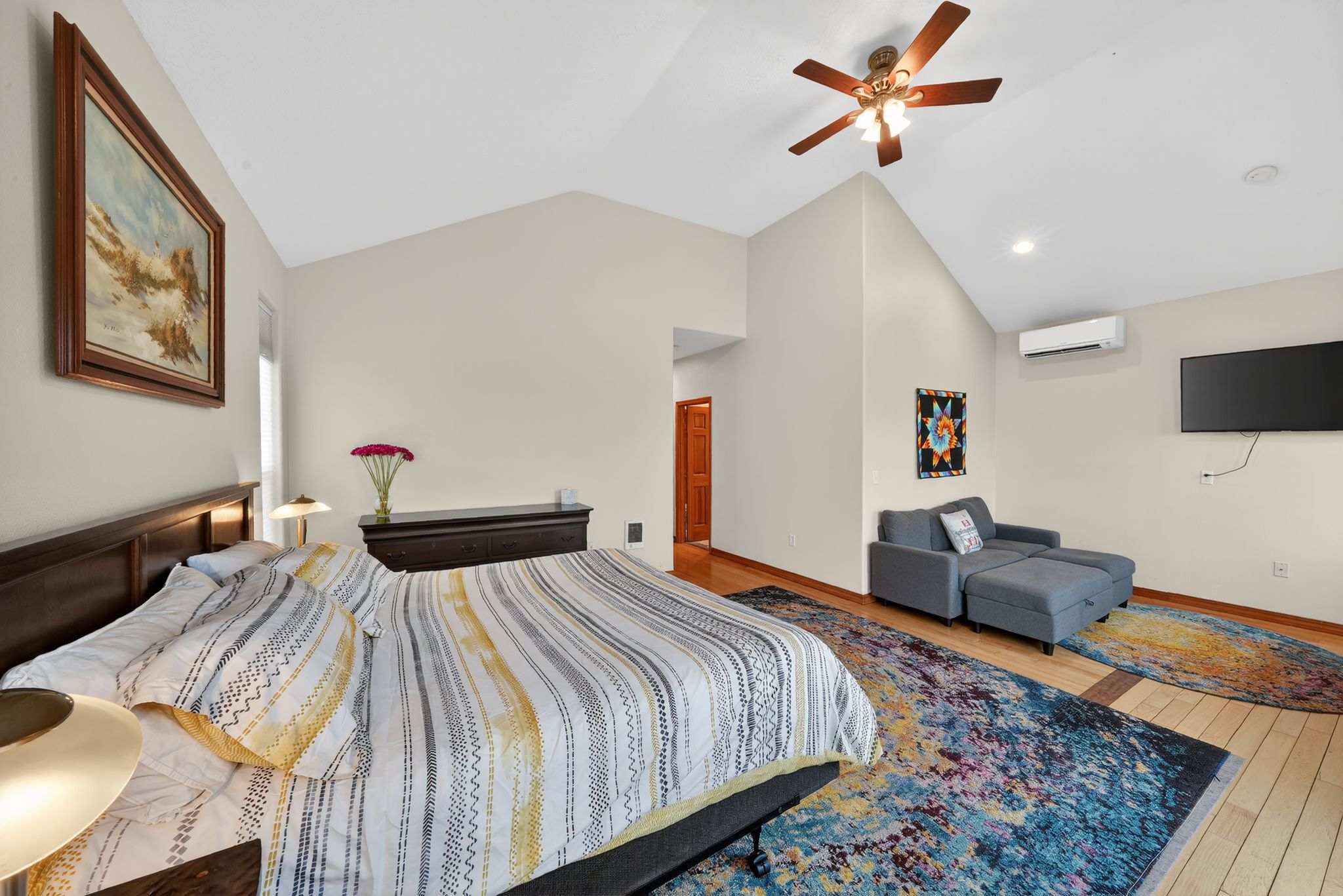 A spacious bedroom with vaulted ceilings and hardwood floors features a bed with a striped comforter and a gray chaise lounge. A colorful abstract rug covers the floor, and a ceiling fan is mounted overhead.