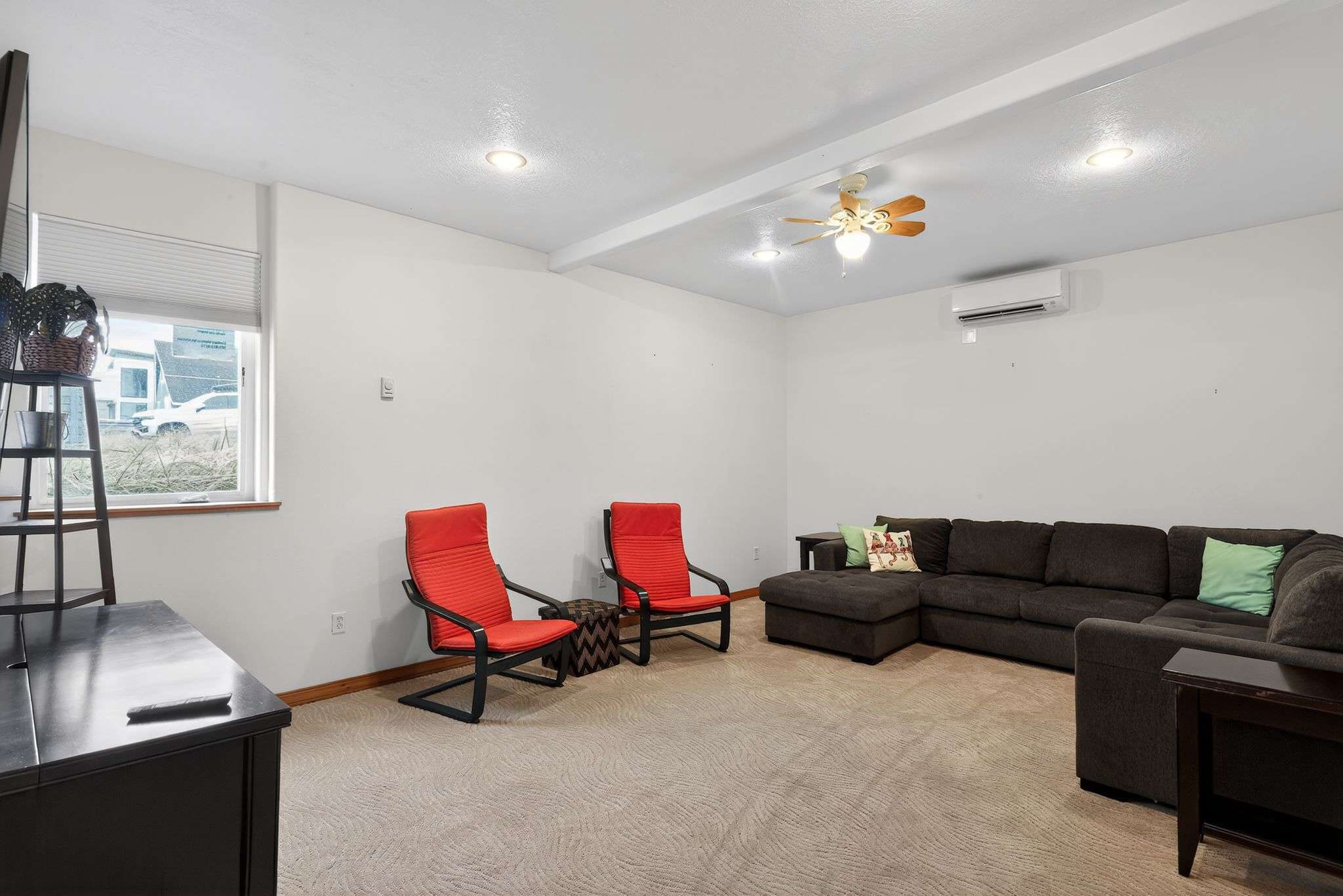 A comfortable living room with light patterned carpet, white walls, a dark sectional sofa, and two modern red chairs, featuring a window and a wall-mounted air conditioning unit, with a ceiling fan for additional comfort.