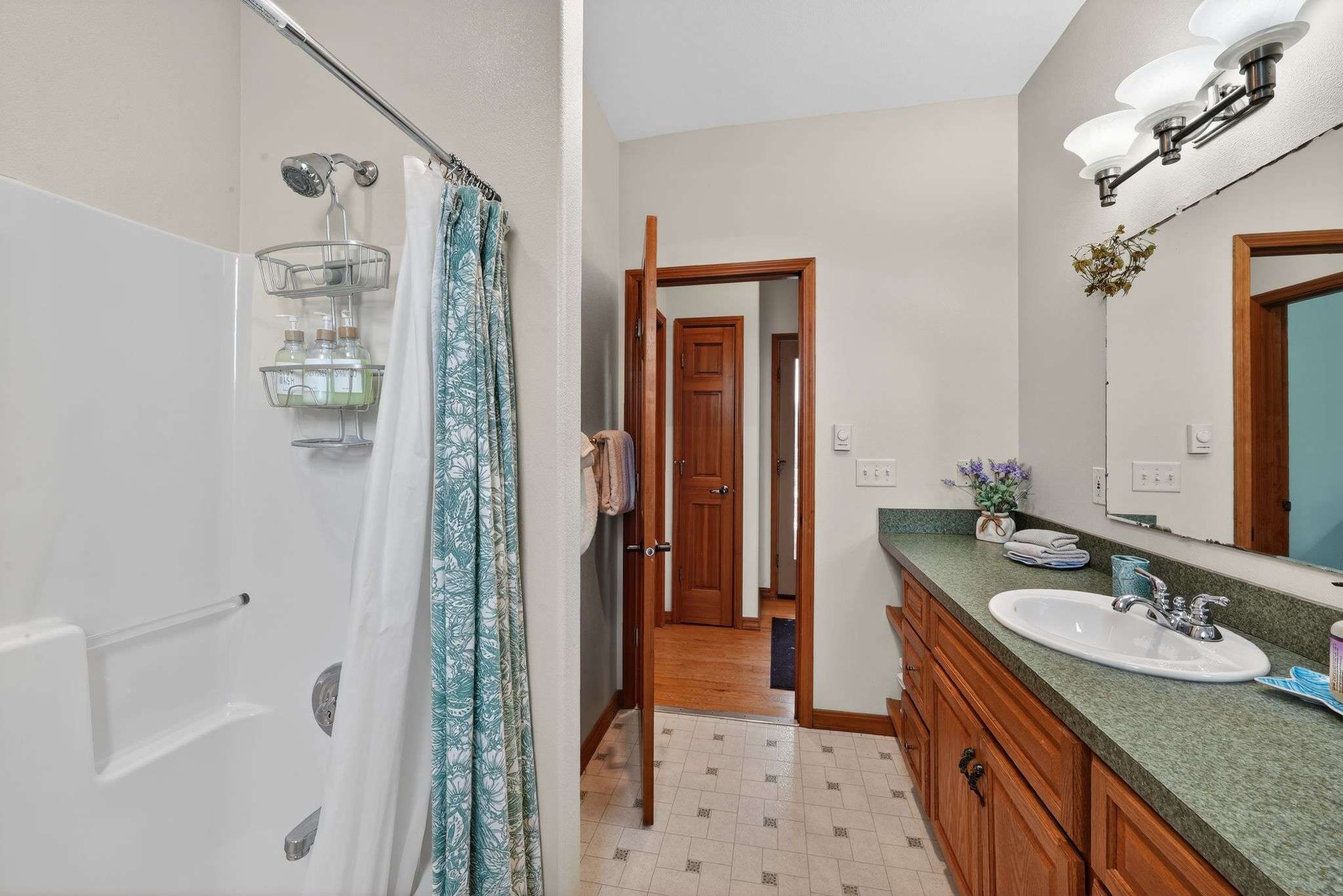 Bathroom with light walls and patterned tiled floors, featuring a single wood vanity with a green countertop, a mirror, and a stand-up shower with a patterned curtain.