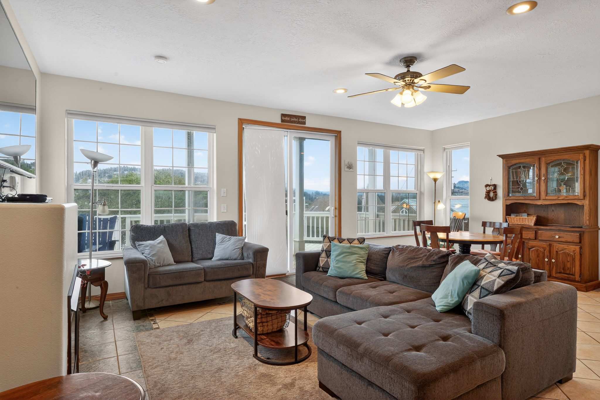 A spacious living room features a large brown sectional sofa and a smaller gray sofa, centered around a wooden coffee table. Multiple large windows offer expansive views of the surrounding landscape, and a ceiling fan hangs overhead.