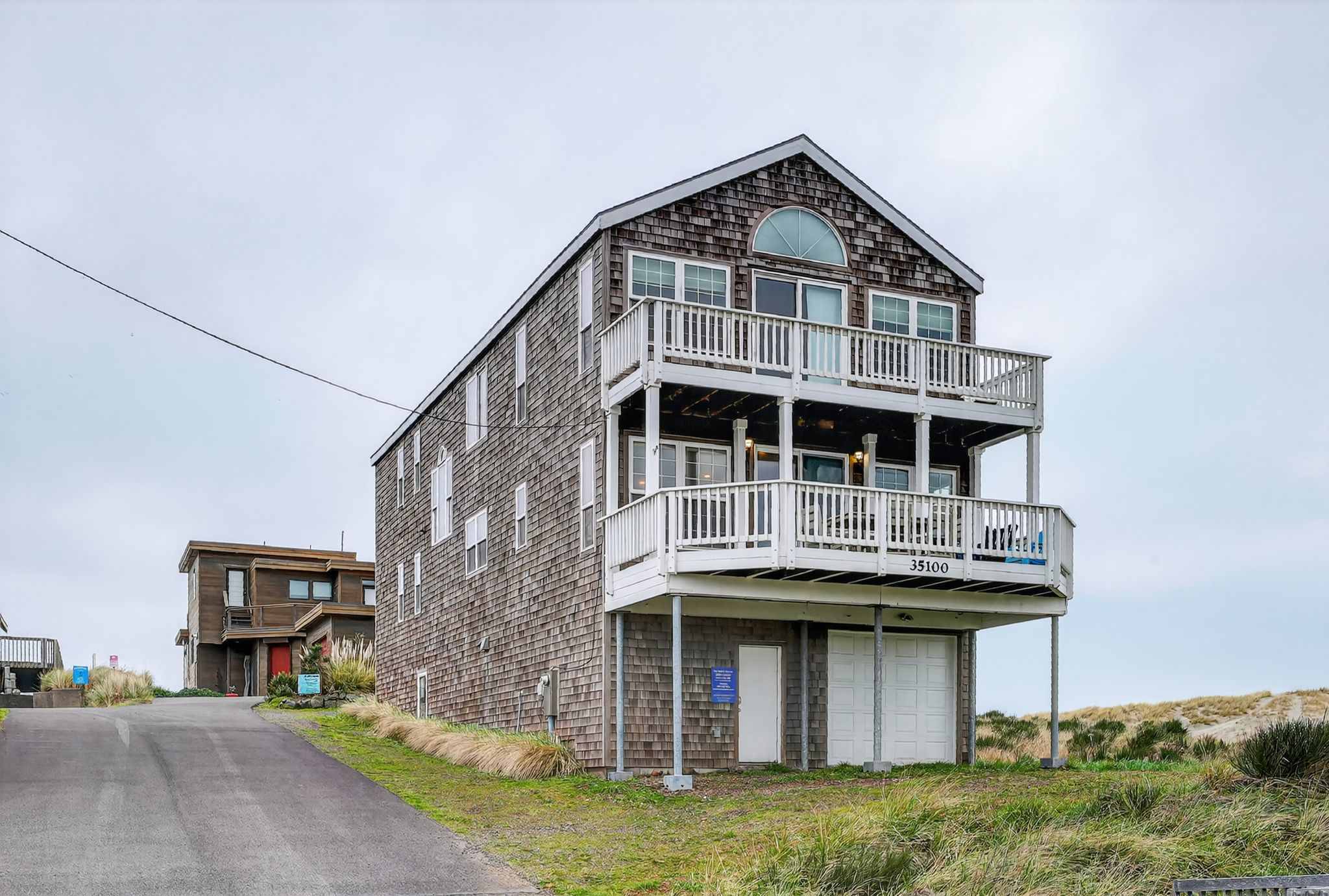 The tall, narrow, three-story house is clad in cedar shakes, featuring multiple balconies with white railings and a ground-level garage. It stands on a grassy dune overlooking the ocean, with the address "35100" visible.