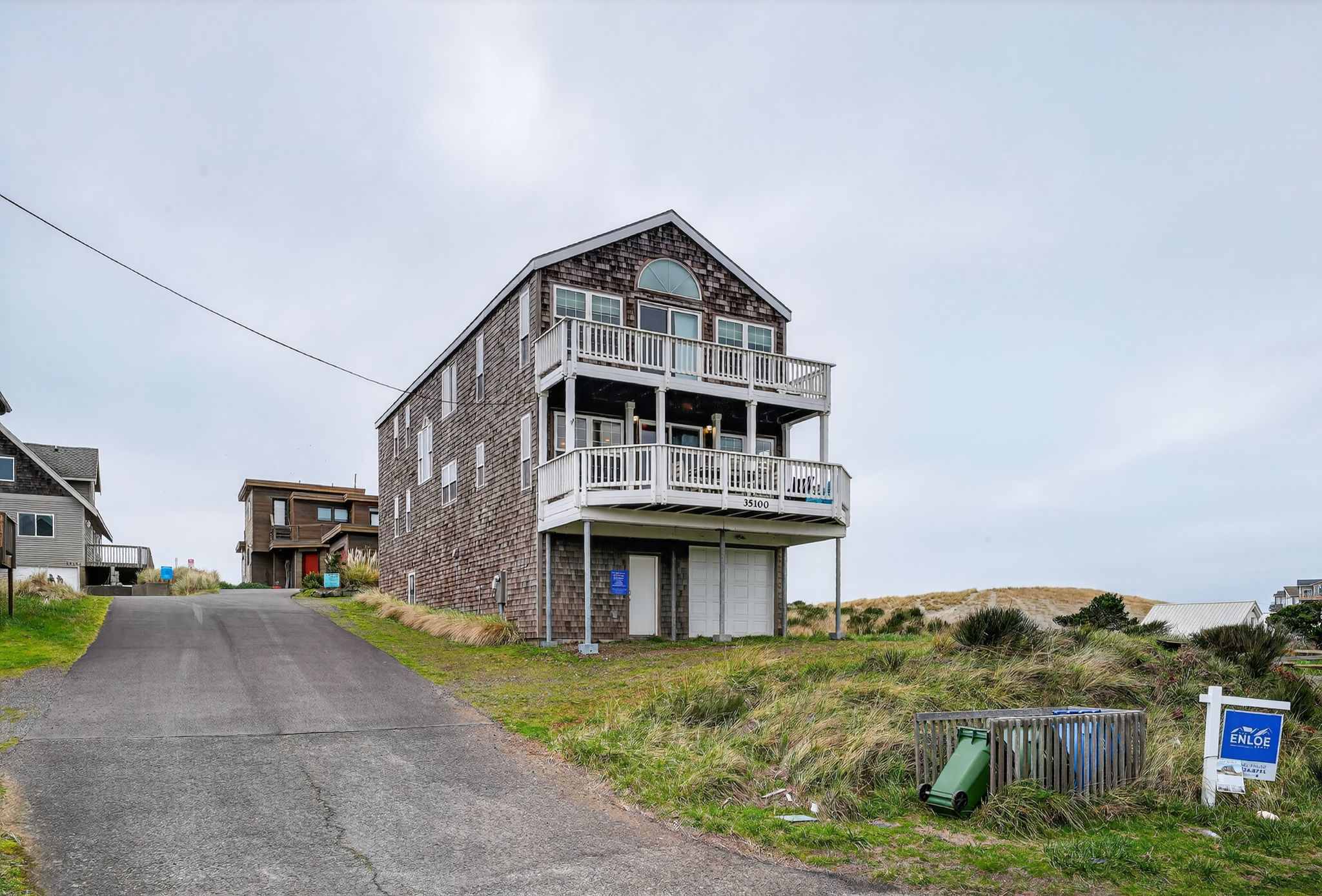The three-story cedar shake house stands on a grassy dune, featuring two balconies with white railings and a ground-level garage. A "For Sale" sign is visible in the foreground, and a neighboring house is in the background.