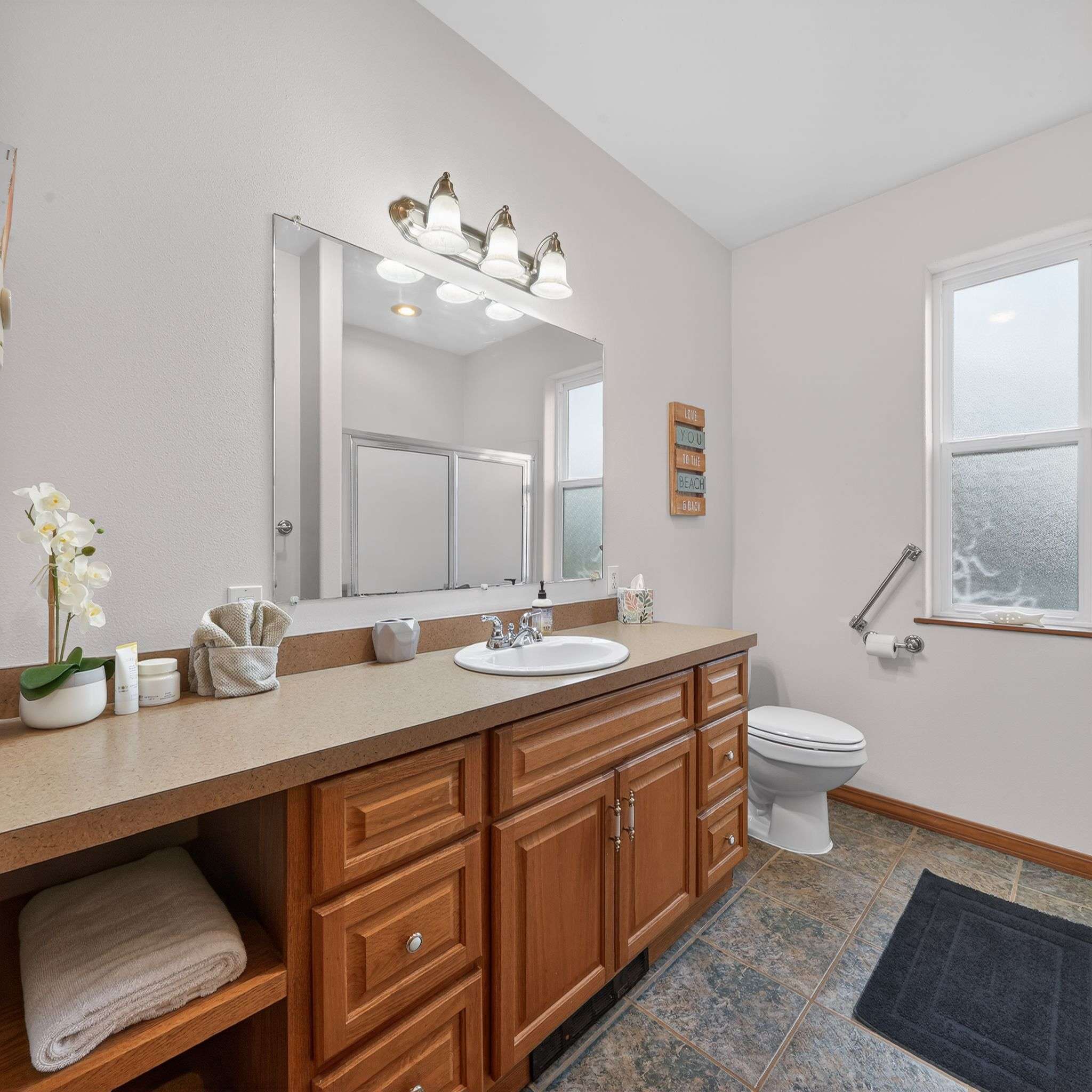 A bright bathroom features a long wooden vanity with a light brown countertop, a single sink, and a large mirror reflecting a shower. A toilet is visible to the right, next to a window with frosted glass and a grab bar.