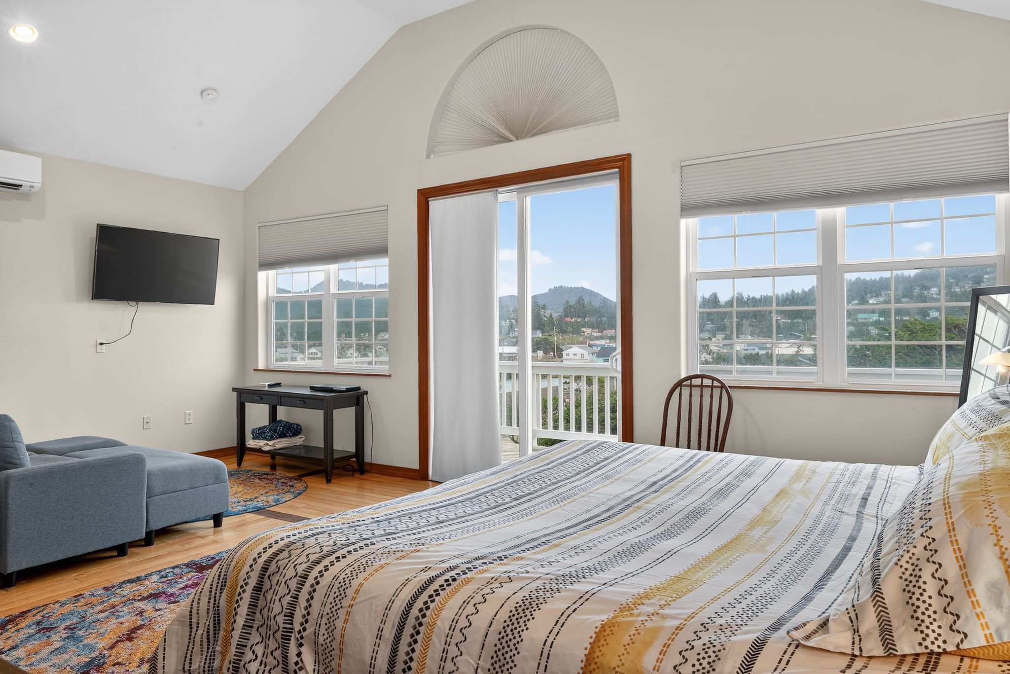 Bright bedroom with a vaulted ceiling, light-colored walls, and hardwood floors, featuring a bed with striped bedding, a blue armchair, and a sliding glass door leading to a balcony with scenic views.