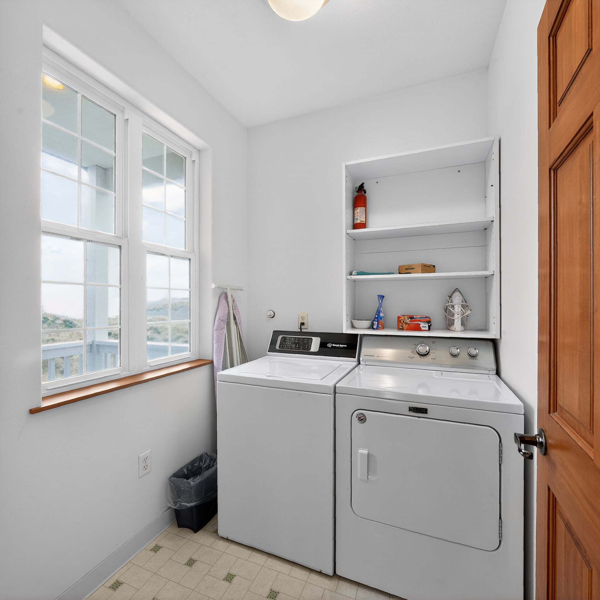 A functional laundry room is equipped with a white top-loading washing machine and a white dryer, situated beneath a window offering an outdoor view. A white shelving unit holds various items, and the floor is covered in light-colored patterned tiles.