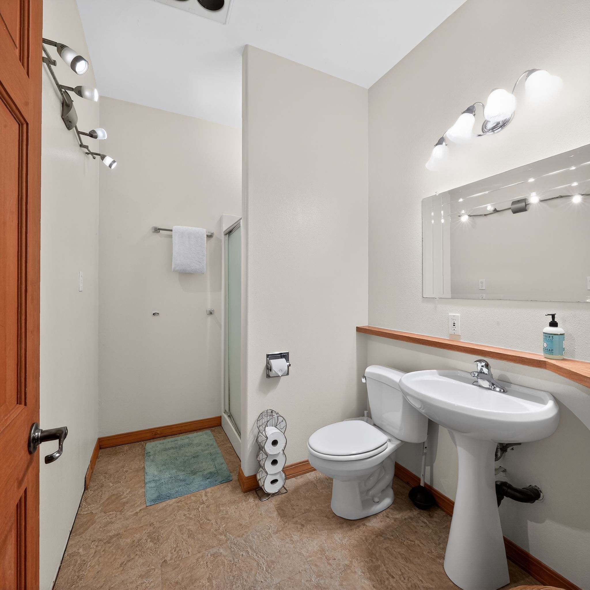 Small bathroom with light walls and patterned linoleum flooring, featuring a white pedestal sink, a toilet, a glass-enclosed stand-up shower, and a wicker basket with towels.