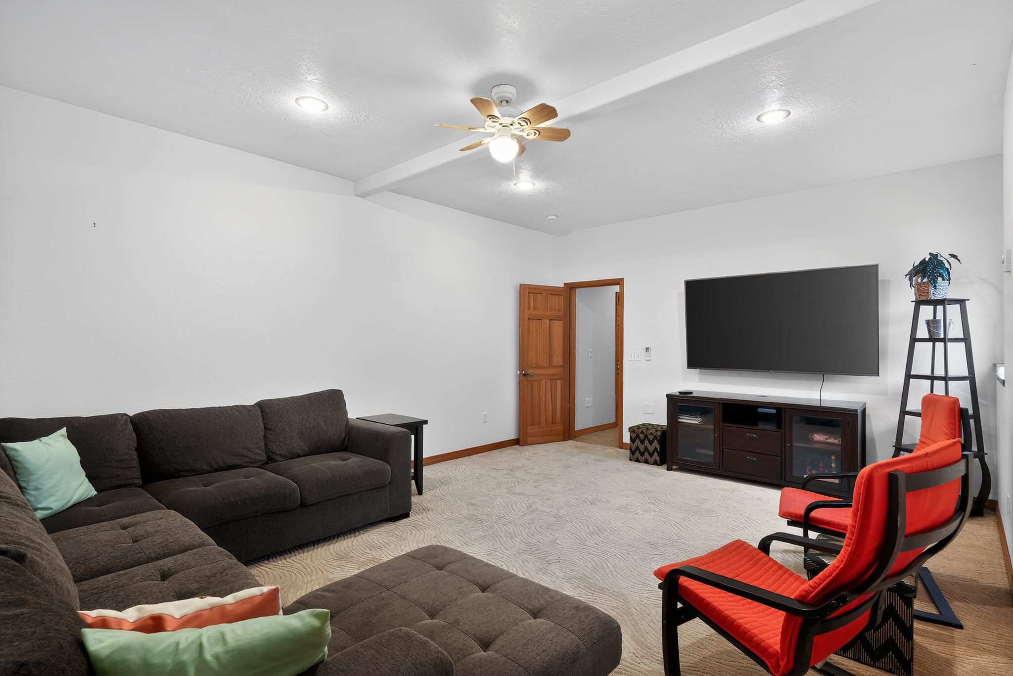 A cozy living room featuring light patterned carpet, white walls, a large dark sectional sofa, two red accent chairs, and a big screen TV on a dark media console, with a ceiling fan overhead.