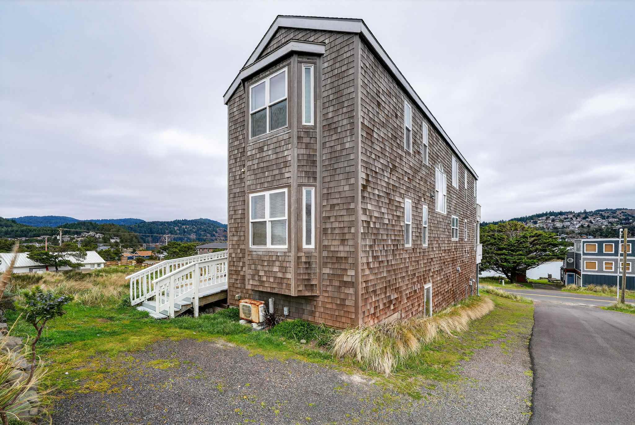 The long side of the cedar shake house is shown, with numerous windows of varying sizes and a wooden ramp leading to the entrance. The property is surrounded by grass and a paved road, with distant hills under a cloudy sky.