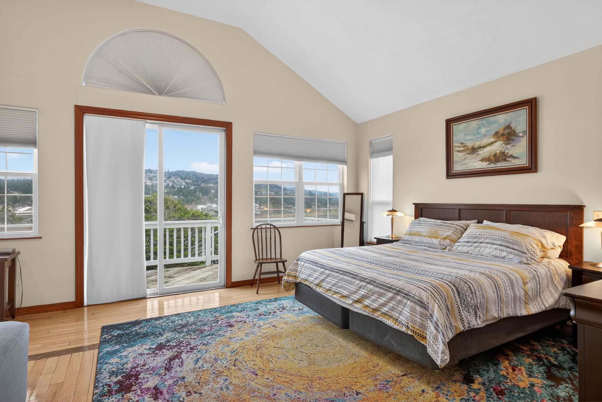 Bright bedroom with a vaulted ceiling, light walls, and hardwood floors, featuring a bed with striped bedding, a wooden chair, and a sliding glass door leading to a balcony with expansive views of hills and houses.